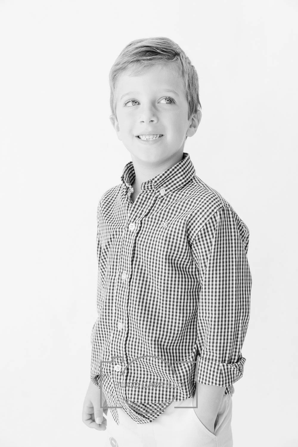 A black and white portrait of a young boy with light-colored hair, wearing a checkered button-up shirt, smiling and looking up and to the side.