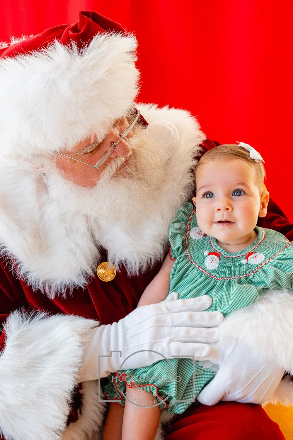 Santa Claus holding a young girl dressed in a green Christmas dress, against a bright red background.