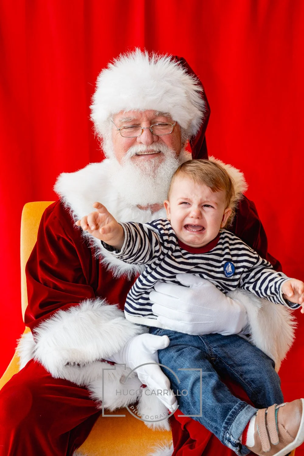 A young child crying and reaching out while sitting on Santa Claus's lap, who is dressed in a traditional red and white Santa suit, with a red curtain background.