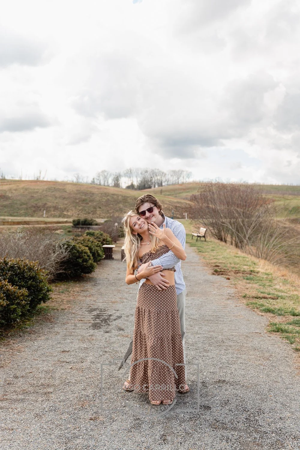 A young couple hugging and smiling on a dirt path in a park, with hills and overcast sky in the background.
