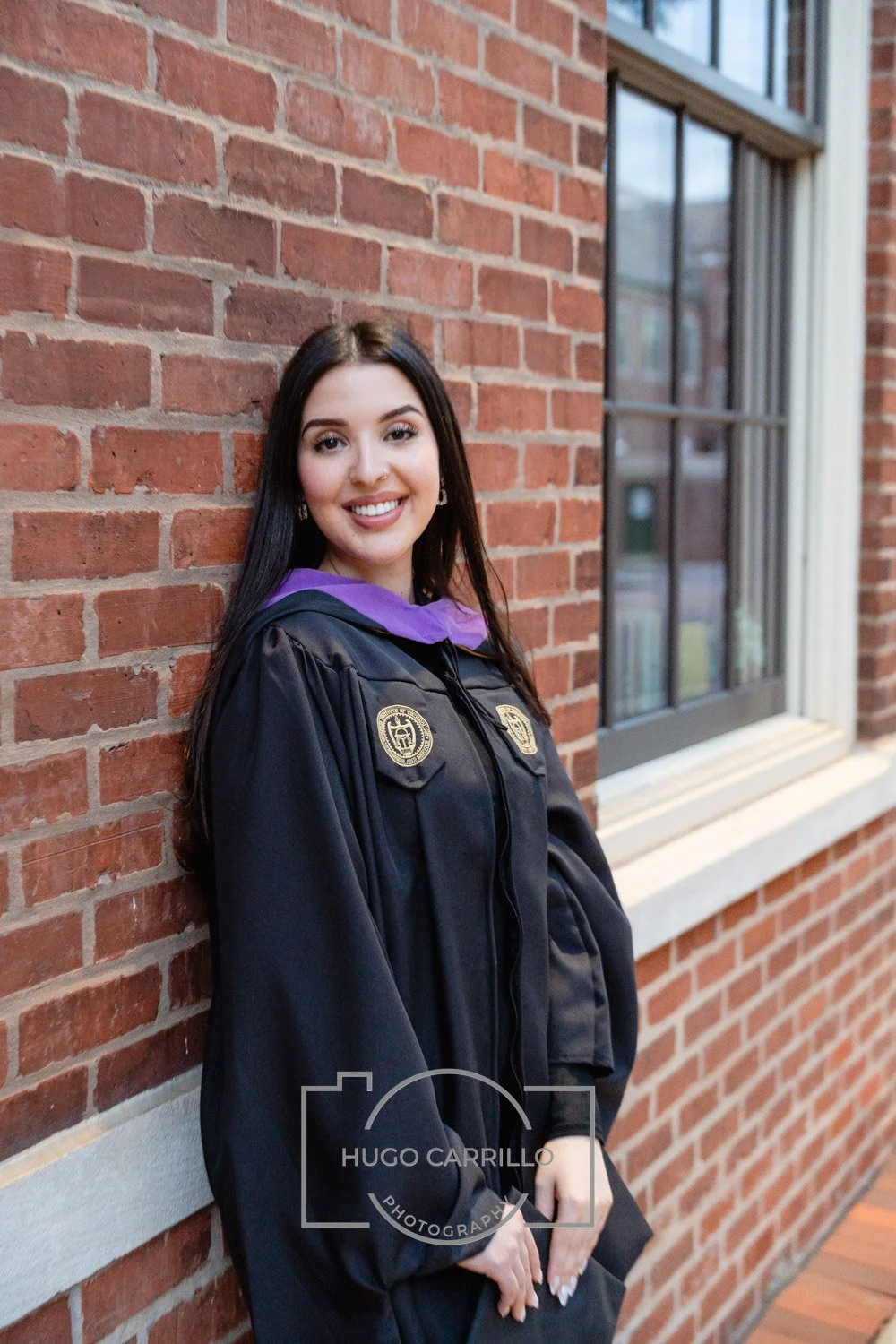 A young woman in a black graduation gown with purple accents, smiling and leaning against a brick wall near a window.