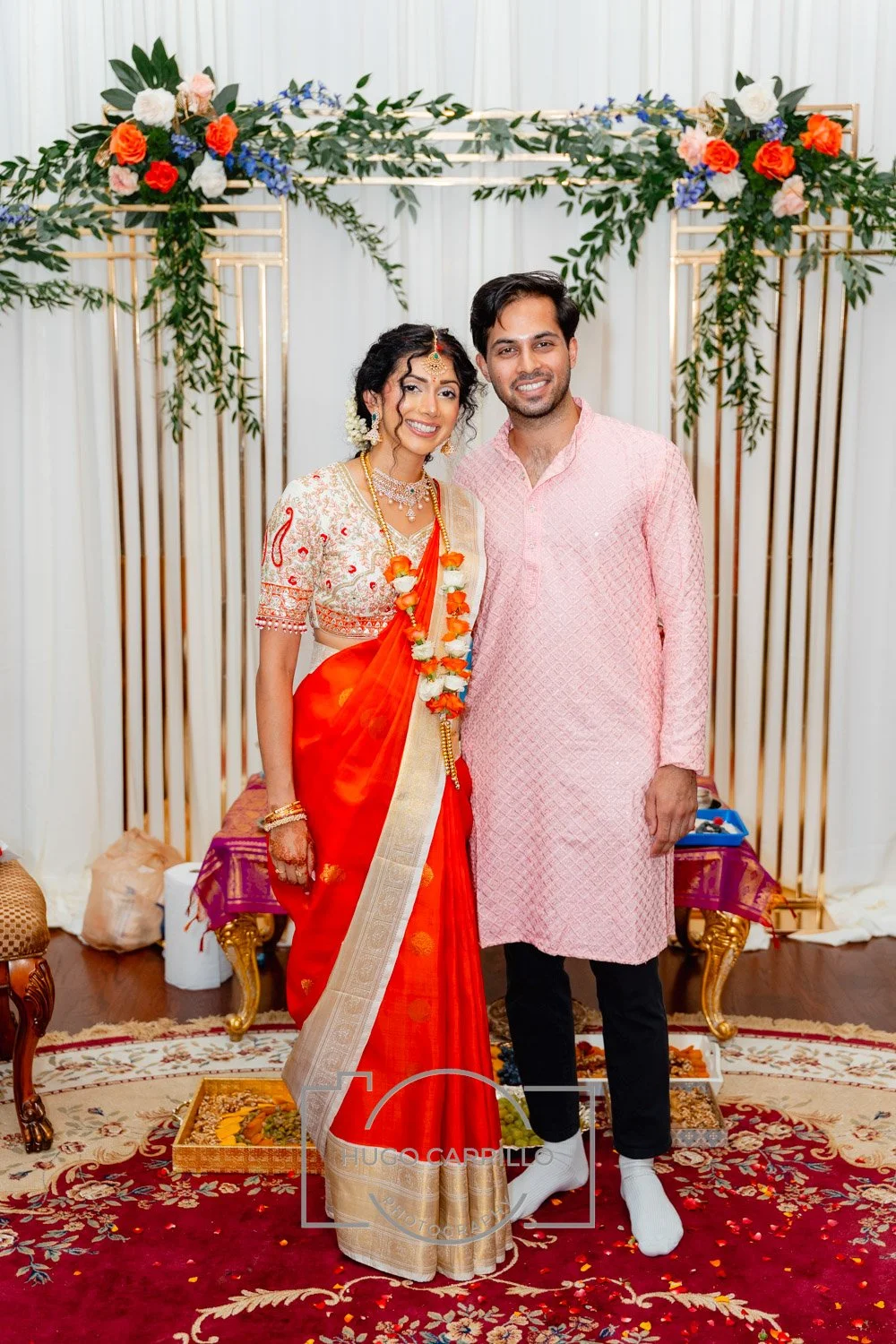 A couple dressed in traditional Indian wedding attire standing together in front of a floral backdrop, smiling for the camera.