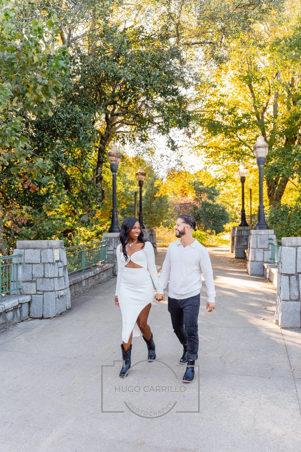 A couple walking hand in hand on a park bridge during sunset, surrounded by green trees with autumn leaves, both smiling and dressed in fashionable white and black clothing.