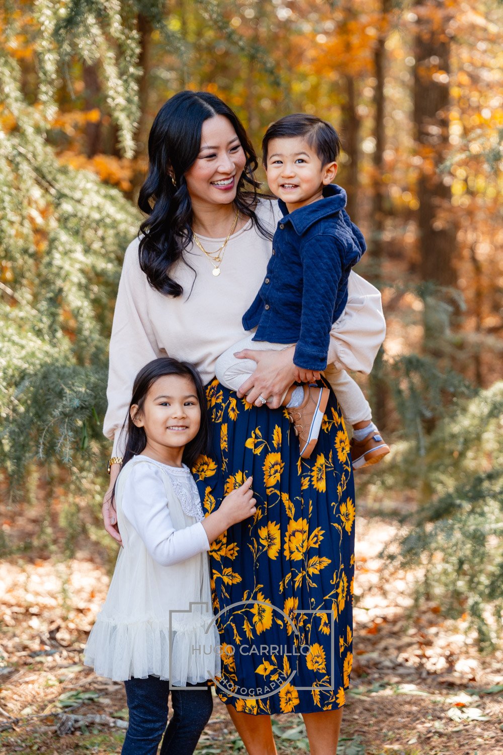 A woman with long dark hair smiling and holding a smiling young boy dressed in a blue jacket. A young girl, smiling and wearing a white dress, stands beside them in an autumnal forest setting.