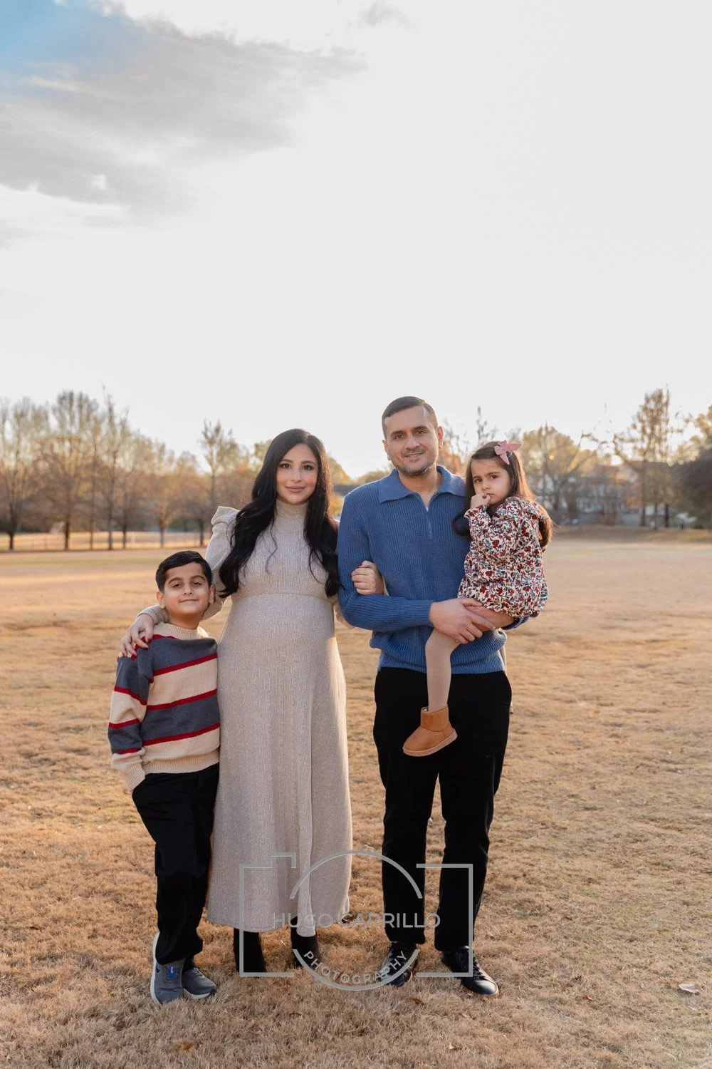 Family of four standing outdoors in a park during fall, with trees and a cloudy sky in the background, smiling at the camera.