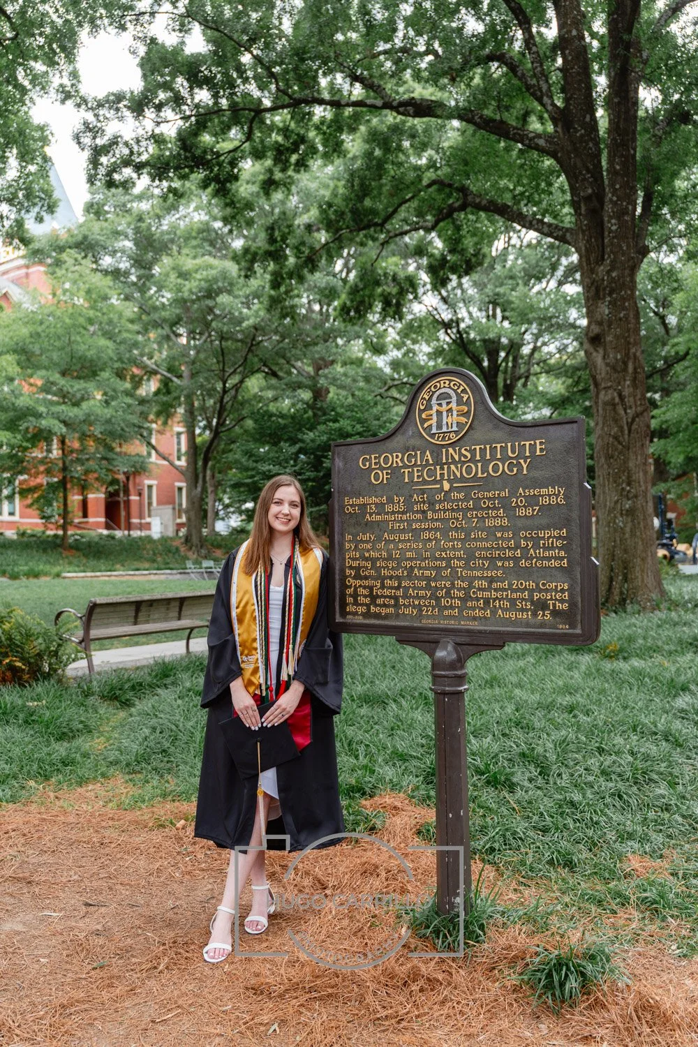 A young woman in a graduation gown with a gold stole and multiple honor cords, holding a diploma cover, standing outdoors in front of a historical marker at Georgia Institute of Technology on a campus with green trees and a brick building in the back