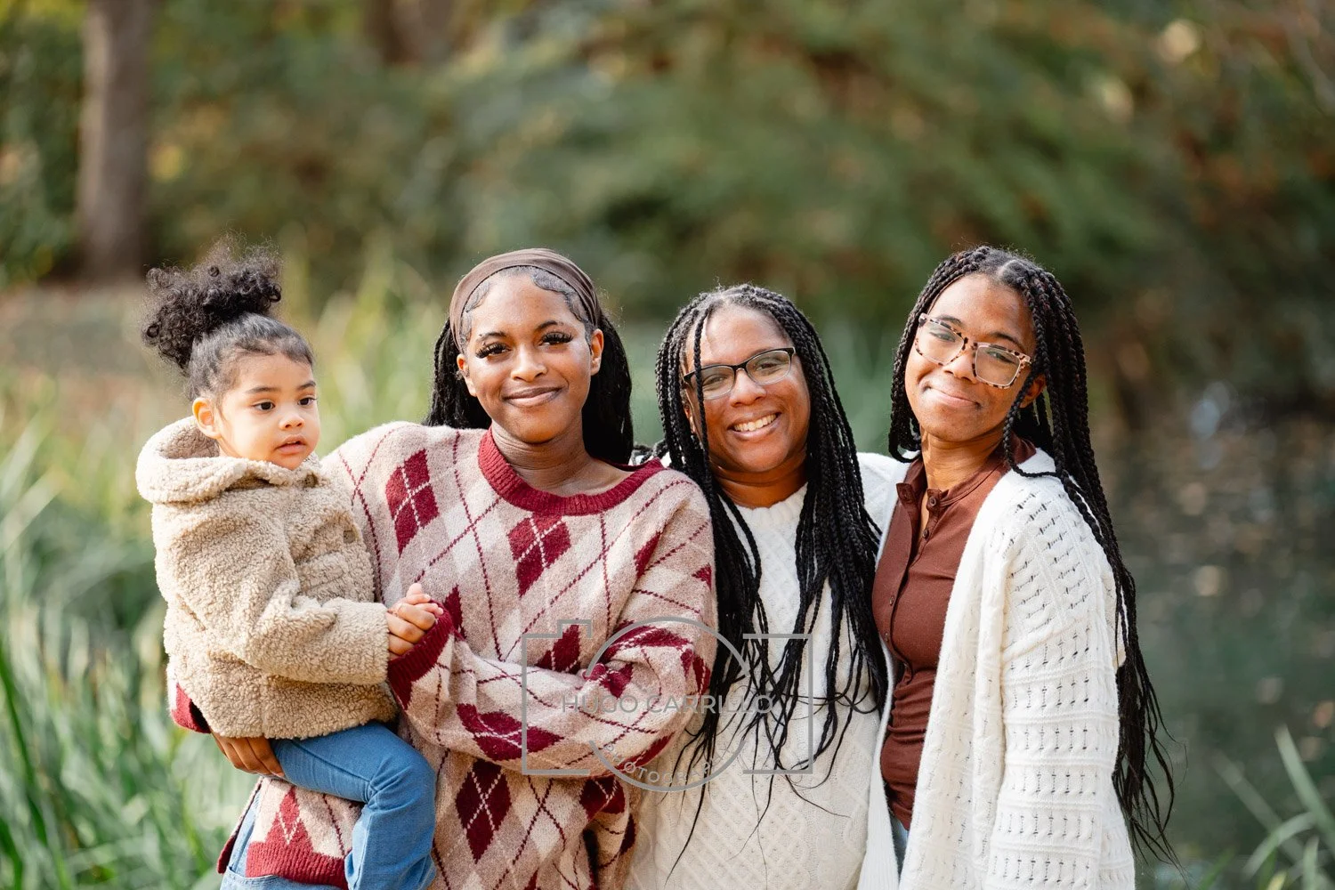 Four women and a young girl standing outdoors near a lake with trees in the background, smiling at the camera.
