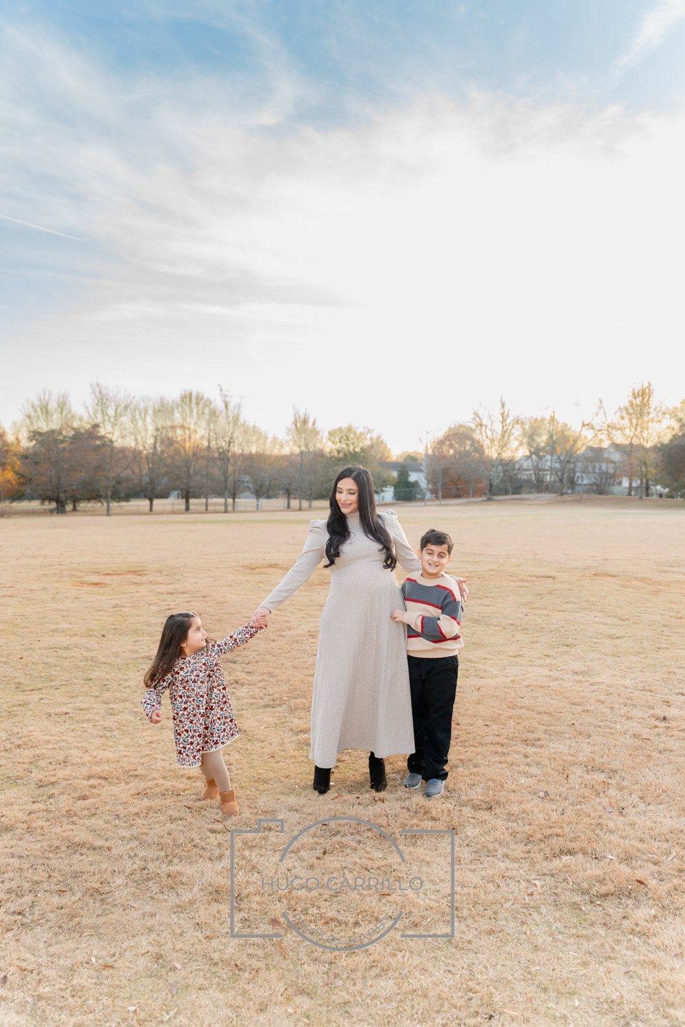 A woman and two children enjoying fall outdoors in a park, holding hands, with trees and houses in the background during sunset.