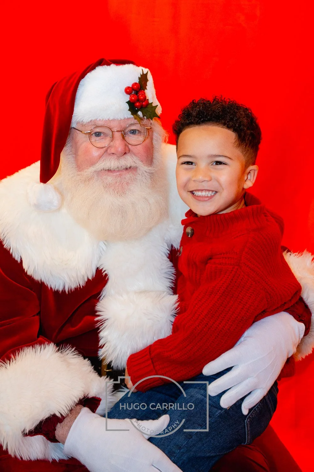 A young boy with curly hair sitting on Santa Claus's lap, both smiling. Santa is dressed in a red suit with white fur trim, a Santa hat with a holly decoration, and glasses. The background is red.