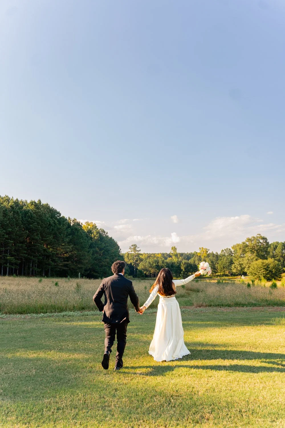 A couple in wedding attire holding hands and walking in a grassy field, with a woman holding a bouquet and a backdrop of trees and a blue sky.