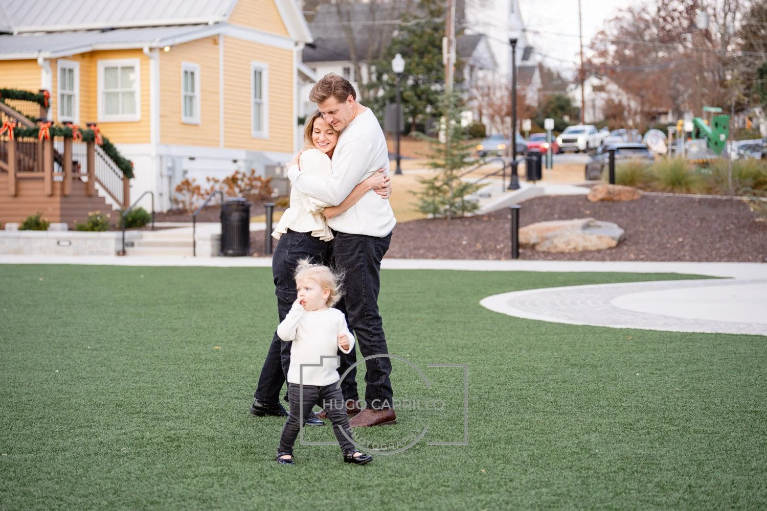 A family of three, a man, woman, and young girl, hugging and standing on a grassy area in a park or playground, with a yellow house and trees in the background.