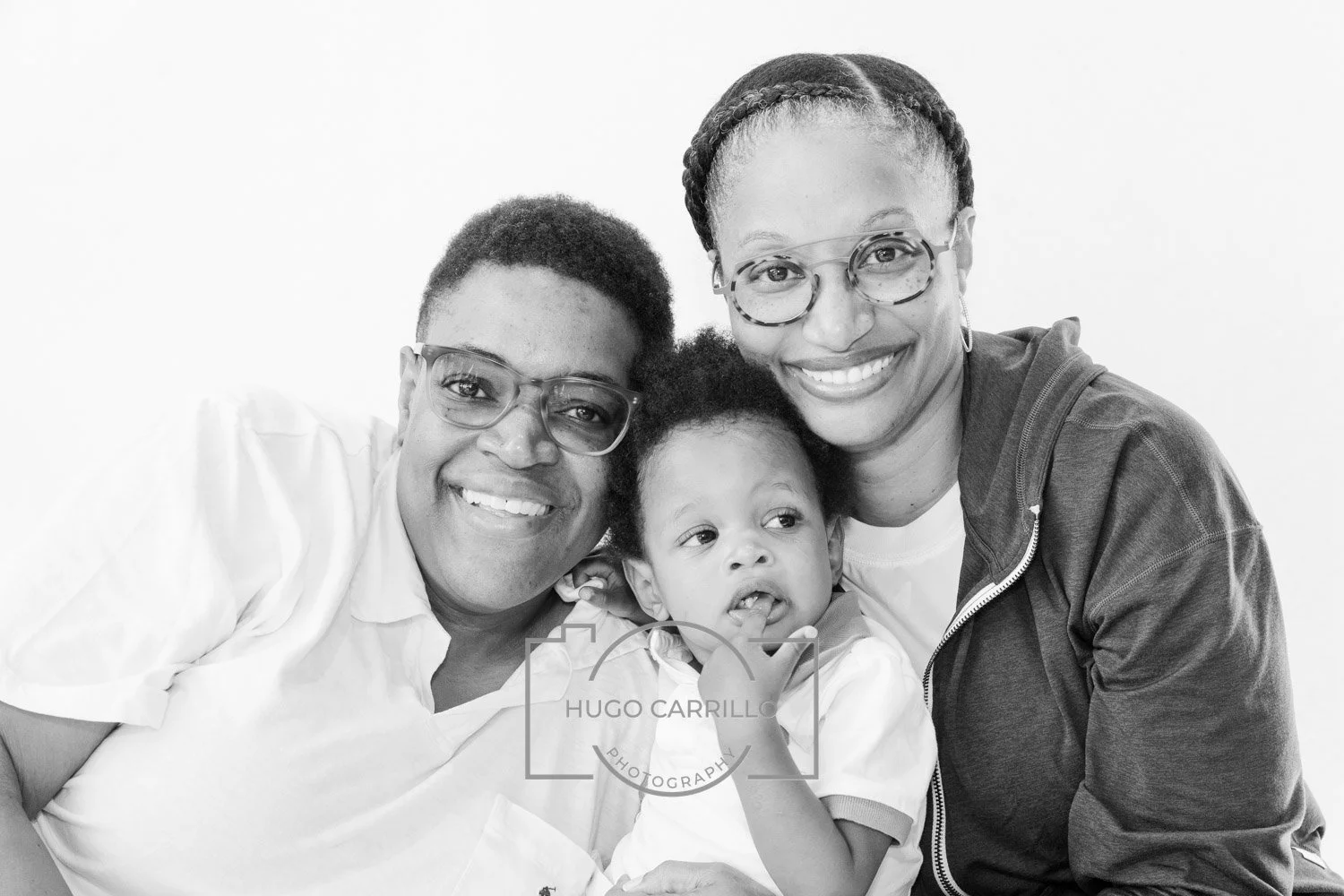 Two smiling women and a young child with curly hair, all wearing glasses, close together in a black and white portrait.