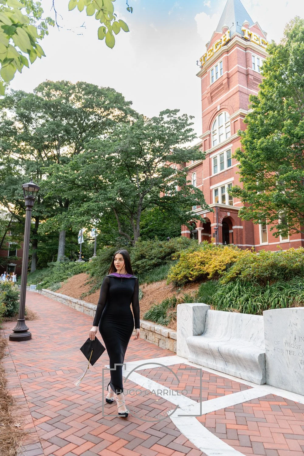 A young woman in a black dress with sheer sleeves and clear heels walking on a brick path with trees, greenery, and a historic red brick building with a clock tower behind her.