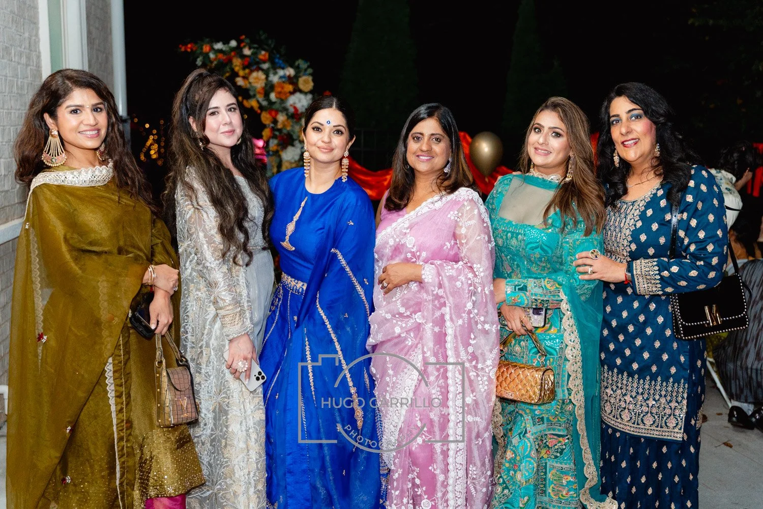 Group of seven women dressed in colorful traditional Indian sarees and outfits, standing outdoors at night, with decorative flowers and balloons in the background.