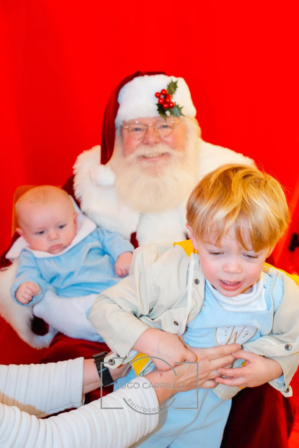 A young boy in a light-colored jacket being surprised by Santa Claus, who is holding a baby, with a red background.