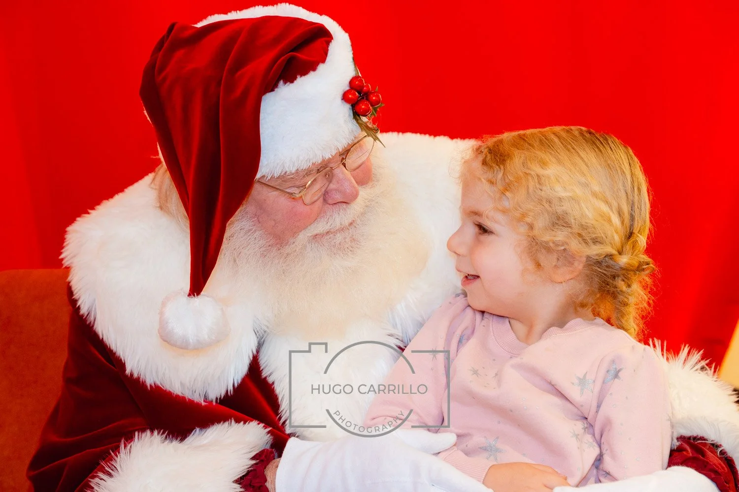 Santa Claus in a red velvet coat with white fur trim, wearing a matching red hat with a white pom-pom and holly decoration, sitting with a young girl with curly blonde hair, against a red background.