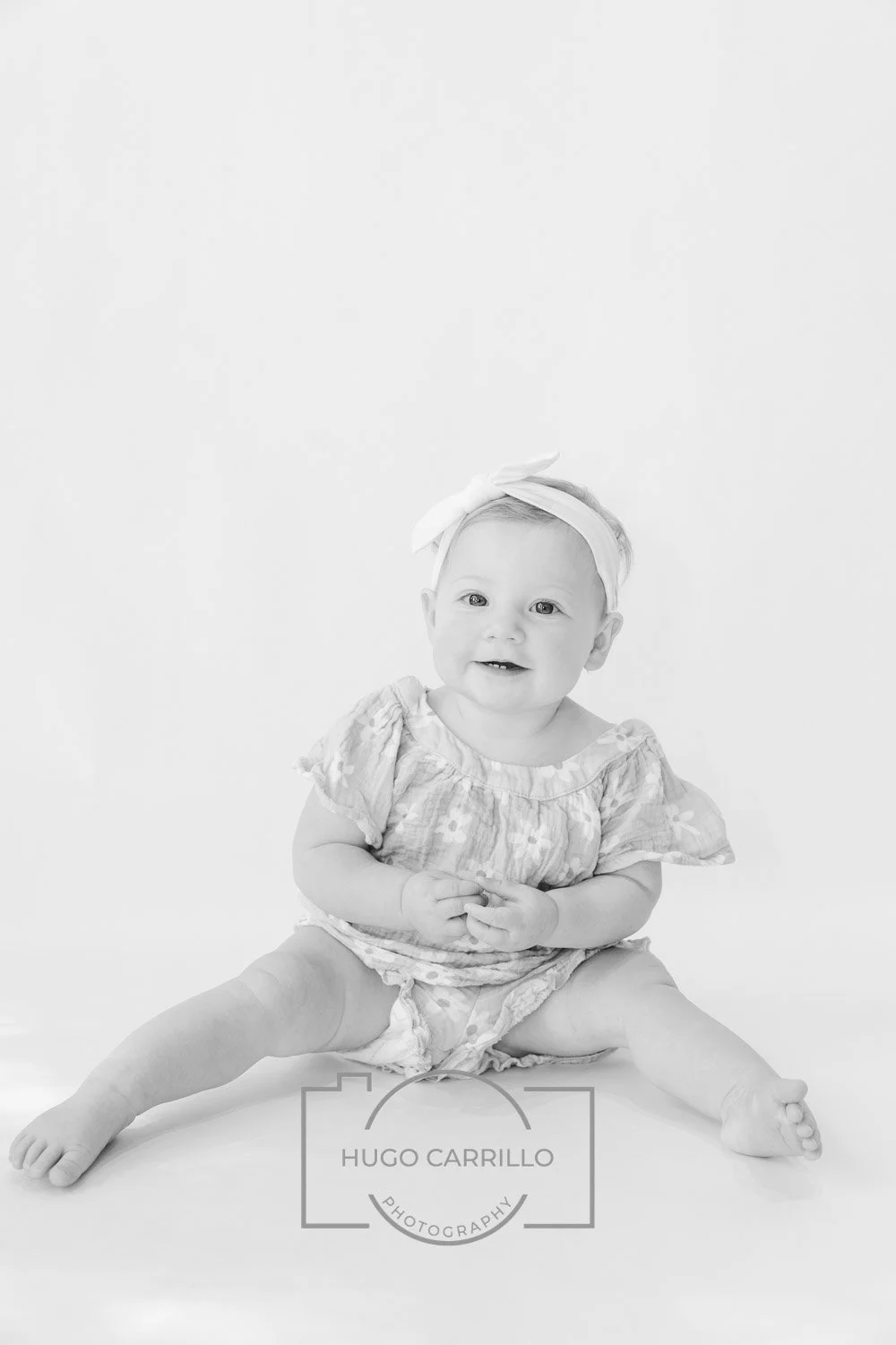 A smiling baby girl with a headband, sitting on the floor with legs spread, in a light patterned dress, against a plain white background.