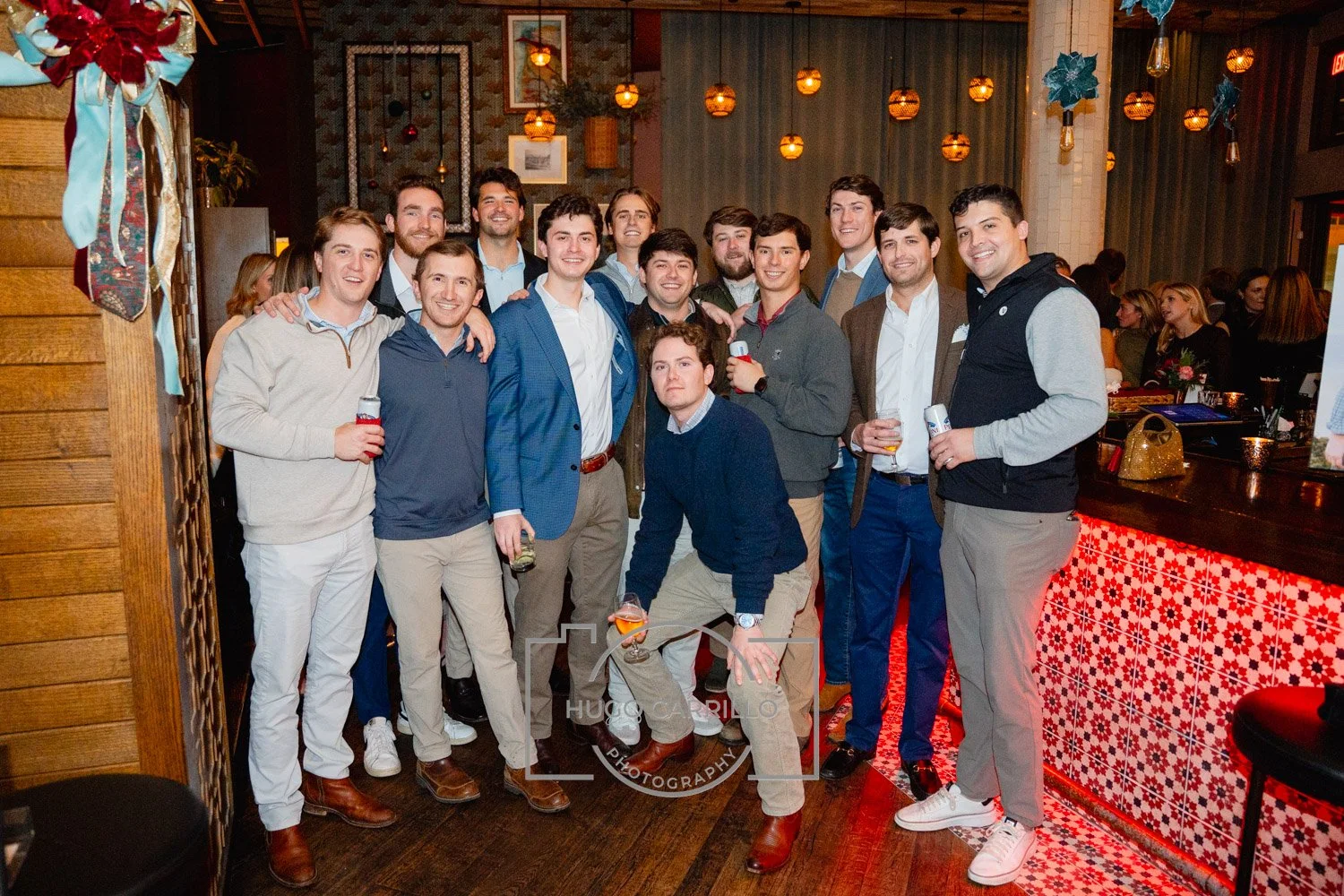 A group of fifteen young men gathered together at a social gathering in a decorated indoor setting, smiling for the camera, with some holding drinks.