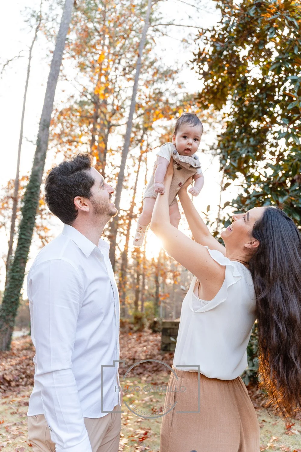 A family of three outdoors during autumn. The woman lifts a baby above her head while the man looks at them. Trees with fall foliage and sunlight in the background.