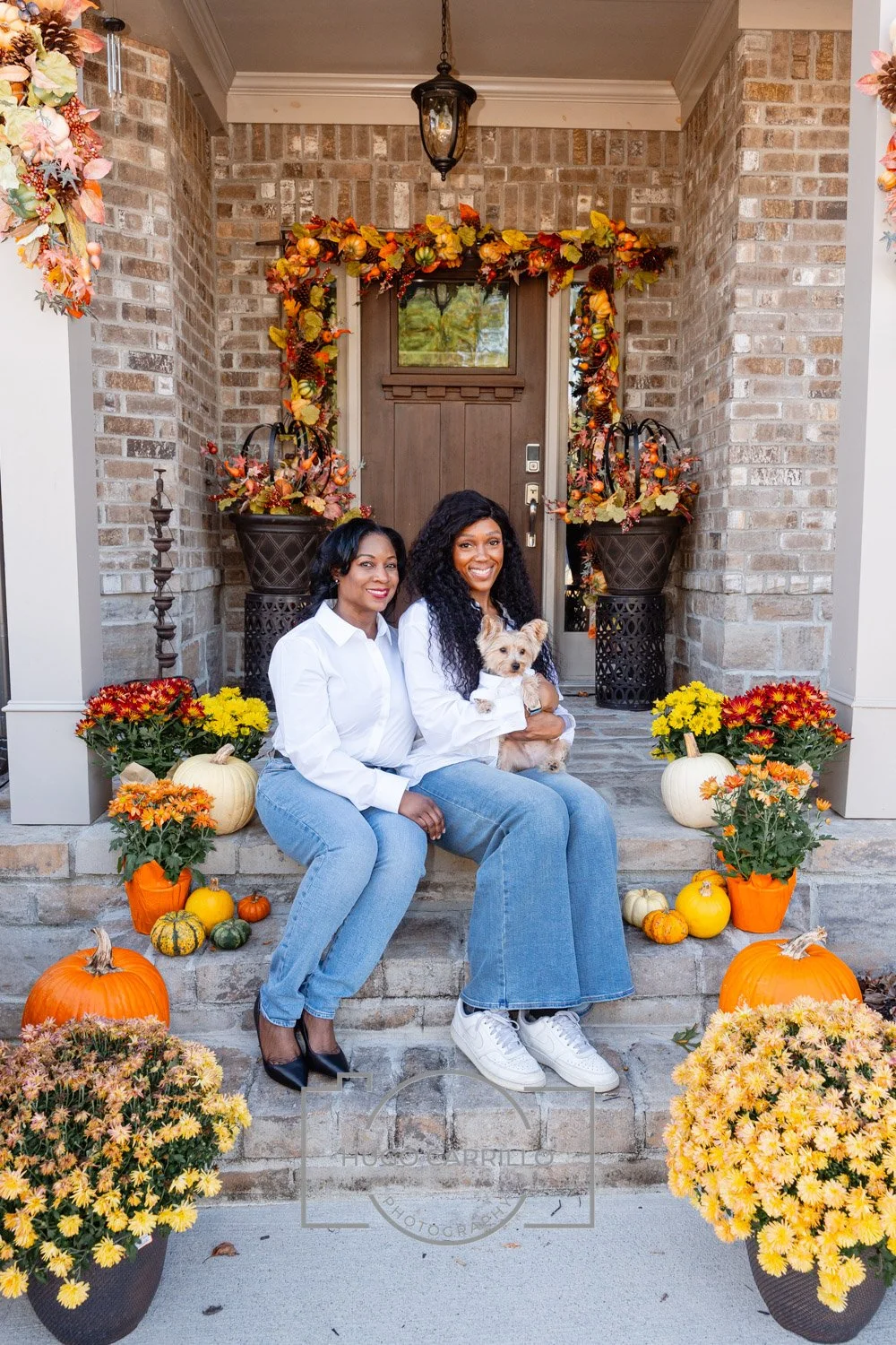 Two women sitting on the front steps of a house decorated for fall, with pumpkins, mums, and autumn leaves, holding a small dog.