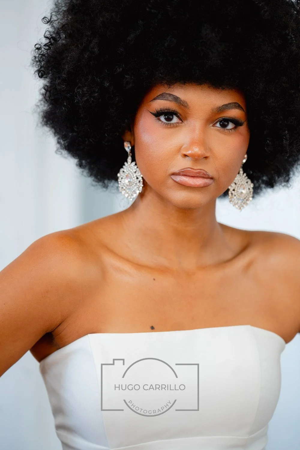 A woman with dark skin and a large, curly afro hairstyle wearing silver earrings and a strapless white dress, looking directly at the camera with a neutral expression.