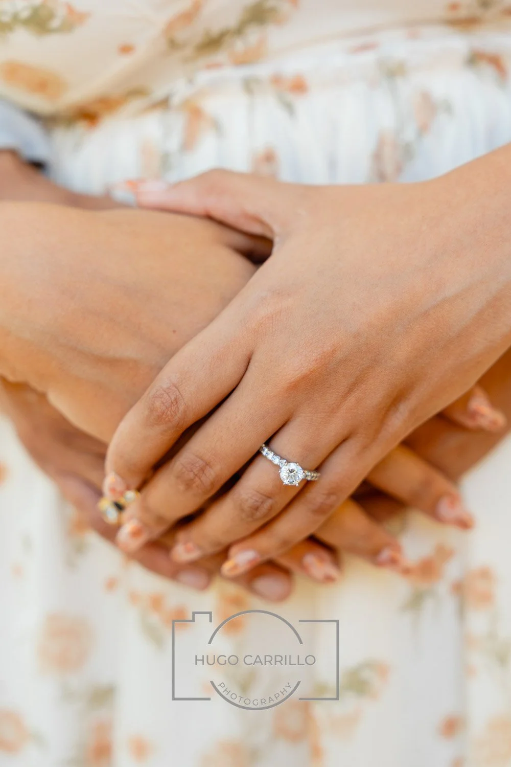 Close-up of a woman's hand with a diamond engagement ring, resting on another person's hand, with a blurred floral dress background.