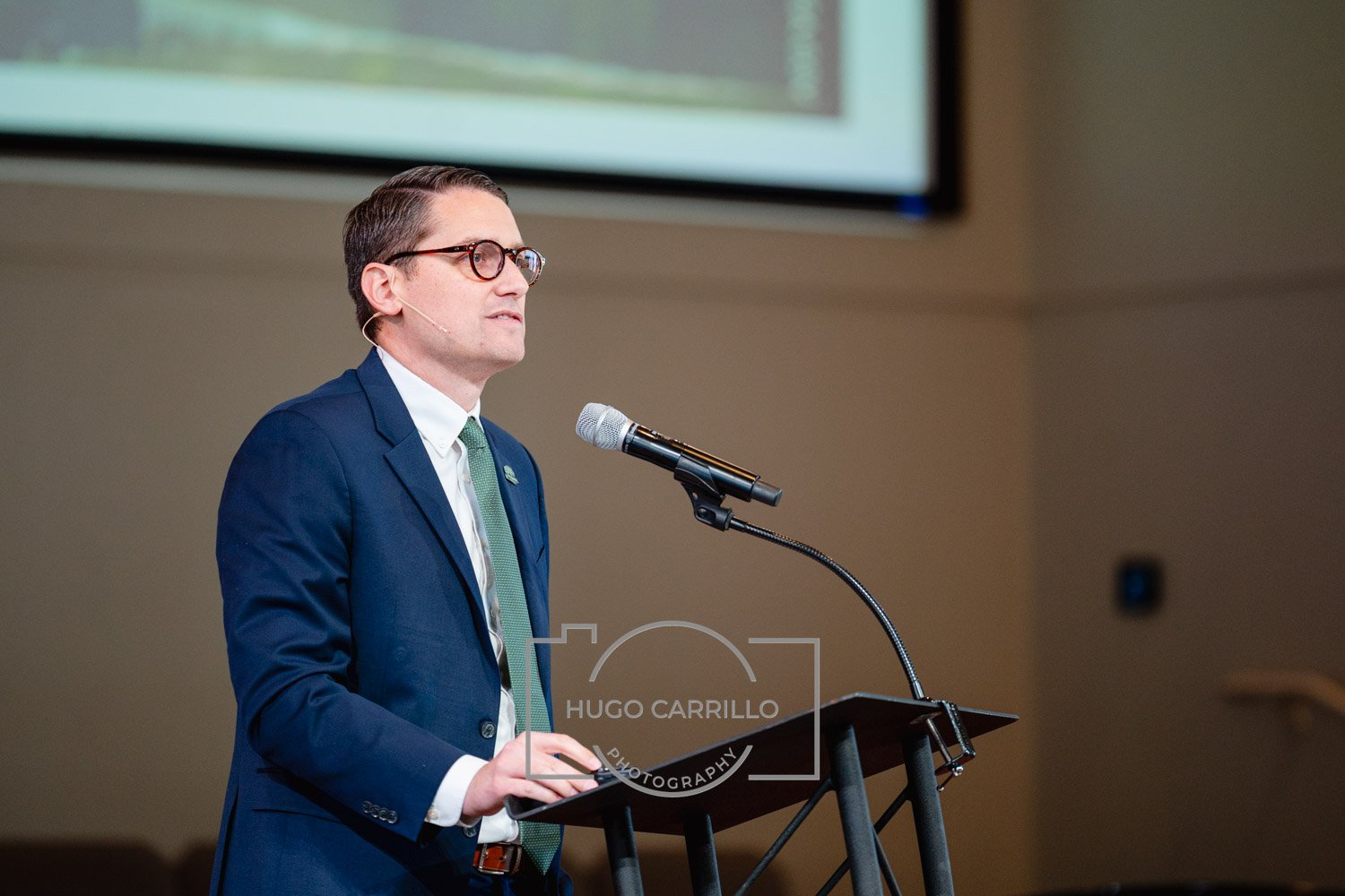 A man in a blue suit, white shirt, and green tie speaking at a podium with a microphone, with a presentation screen behind him.