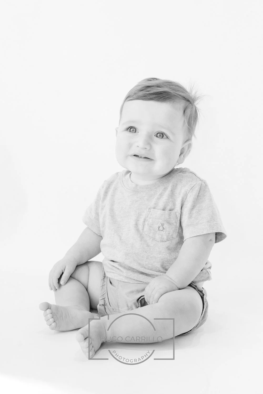 Black and white photo of a toddler sitting on the floor, looking up with a curious expression, wearing a T-shirt and shorts.