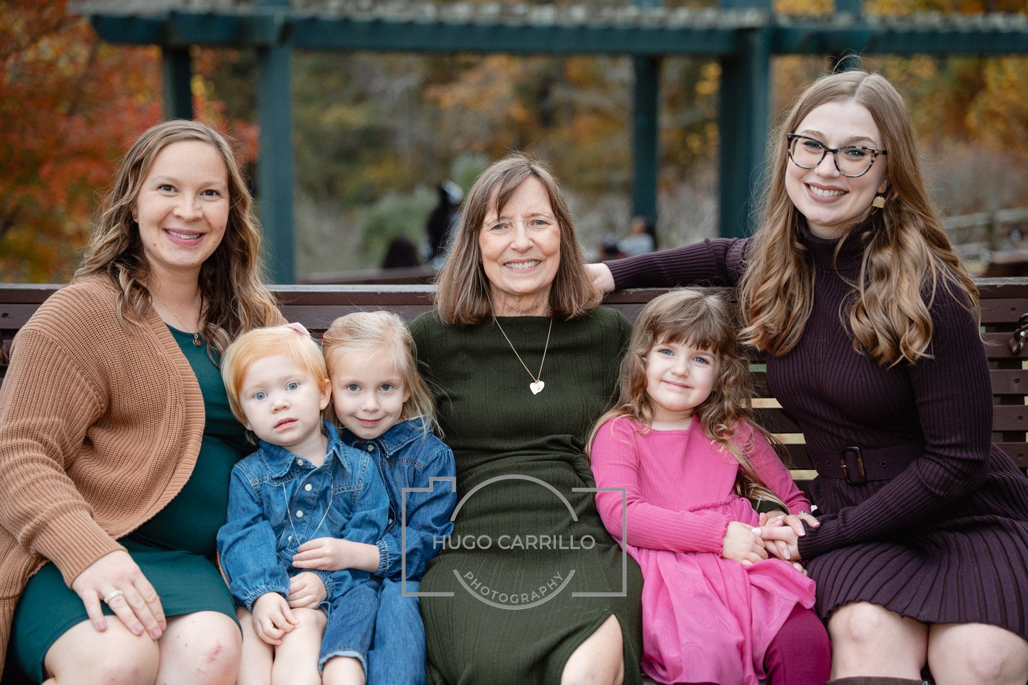 Group of six people, including three women and three children, sitting on a park bench outdoors during autumn. The women are smiling, and the children are sitting between them. The background features autumn trees and a bridge.