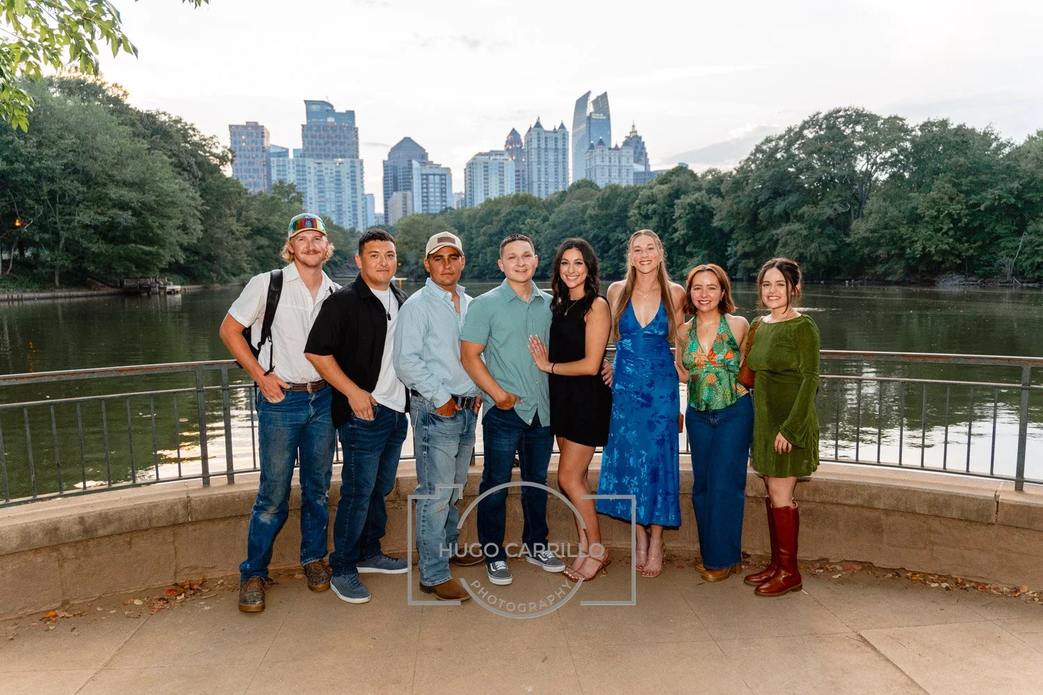 Group of eight friends standing on a riverwalk with city skyline in background, smiling at the camera.