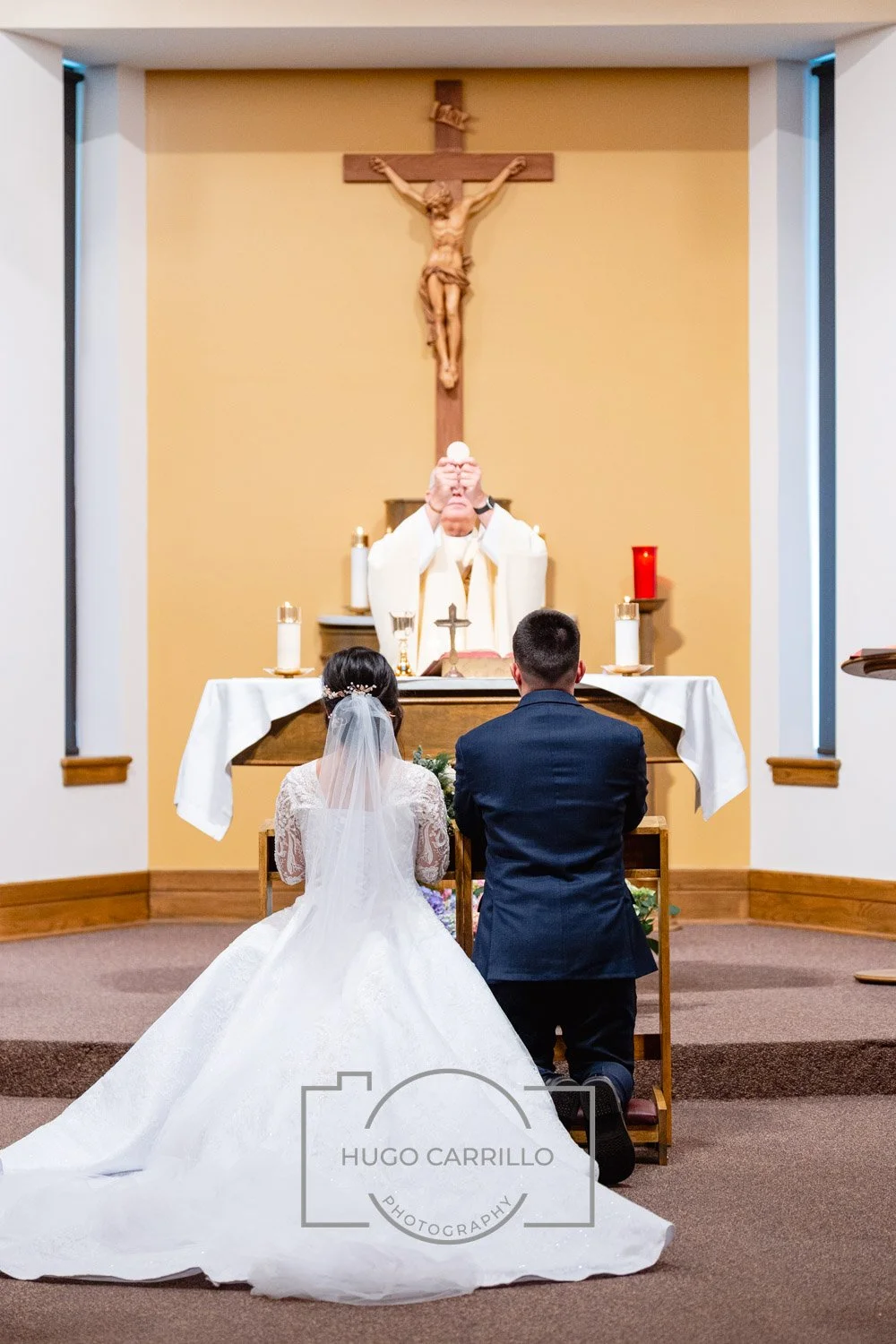 A bride and groom kneeling at an altar during a Catholic wedding ceremony, with a priest holding a Eucharist wafer, in front of a crucifix behind the altar.
