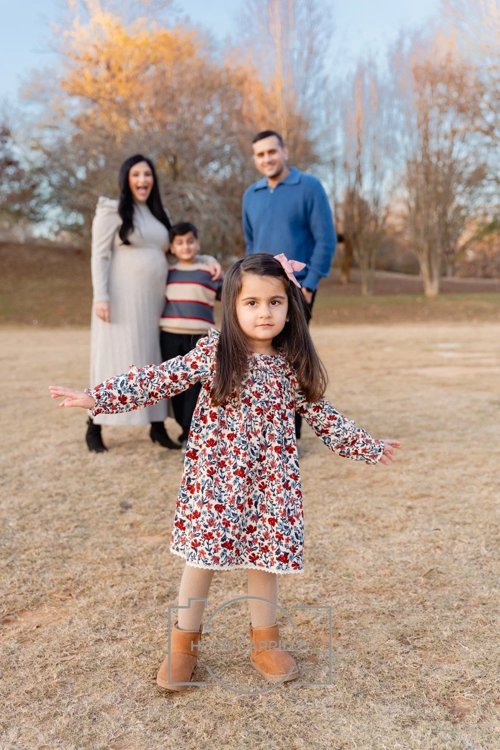 A young girl with long dark hair, wearing a floral dress, tan boots, and a pink bow in her hair, stands in the foreground with her arms outstretched. In the background, a family of four, including a pregnant woman, a man, and a boy, stands outdoors o