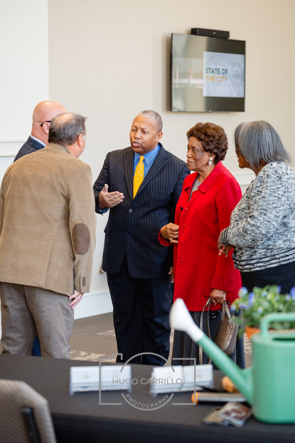A group of five people, three women and two men, engaged in a conversation at an indoor event. One man is speaking while the others listen attentively. They are dressed in business casual and formal attire, with a TV screen displaying a presentation 