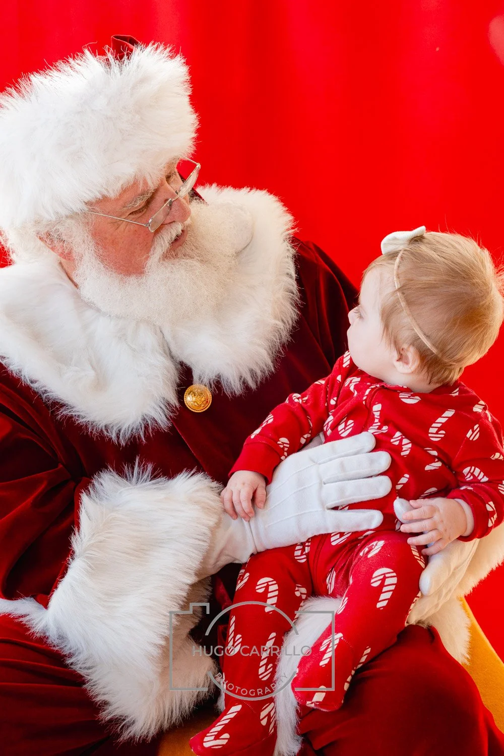 Santa Claus in a red suit with white fur trim holding a young girl in red Christmas pajamas with candy cane patterns, against a red background.