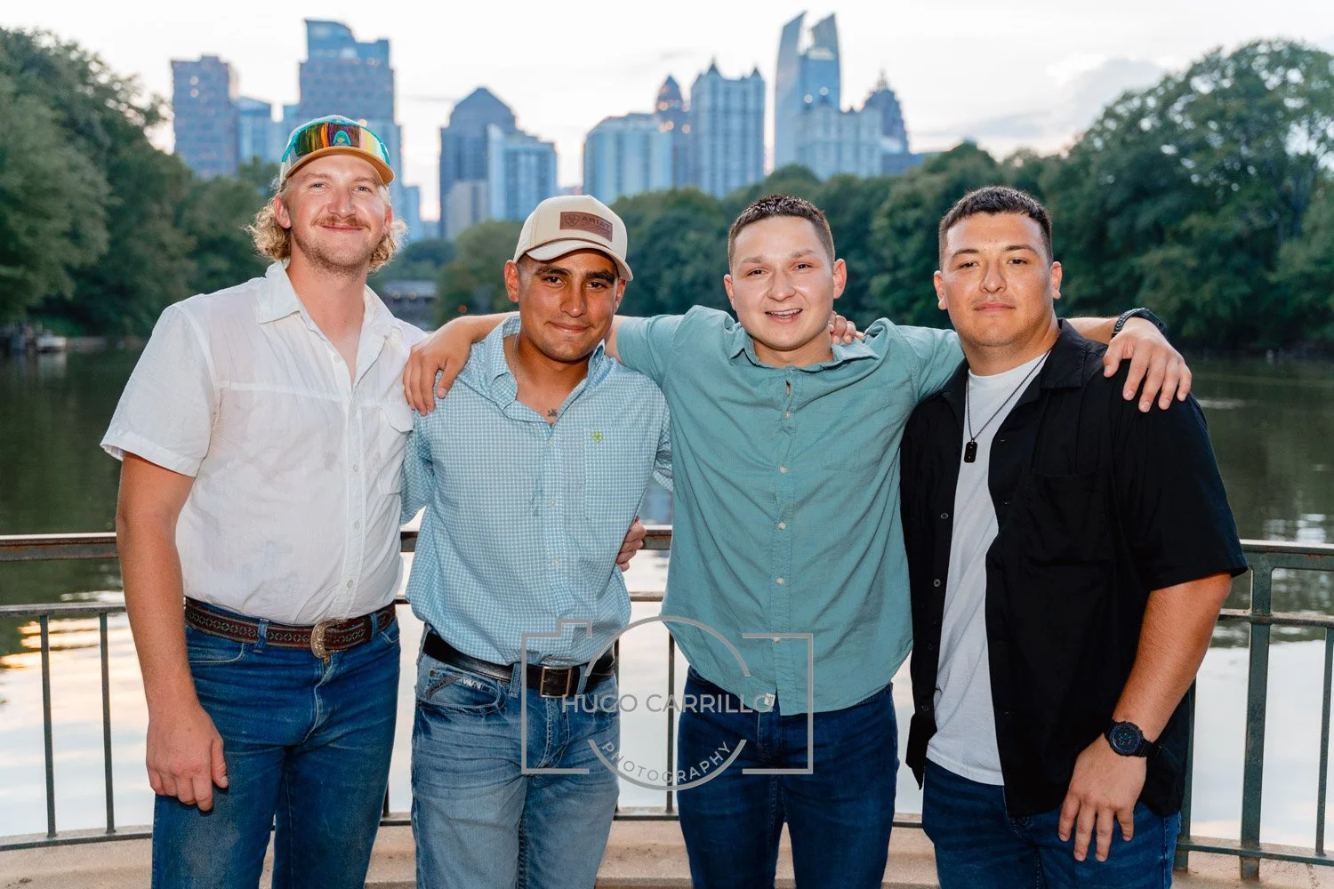 Four men standing arm in arm on a bridge near a river with a city skyline in the background at dusk.