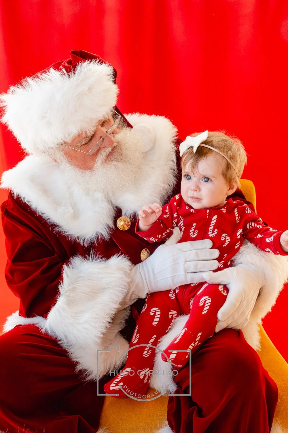 Santa Claus wearing a red suit with white fur trim holding a young girl dressed in red Christmas pajamas with white candy canes and a white bow in her hair, sitting on Santa's lap against a red background.