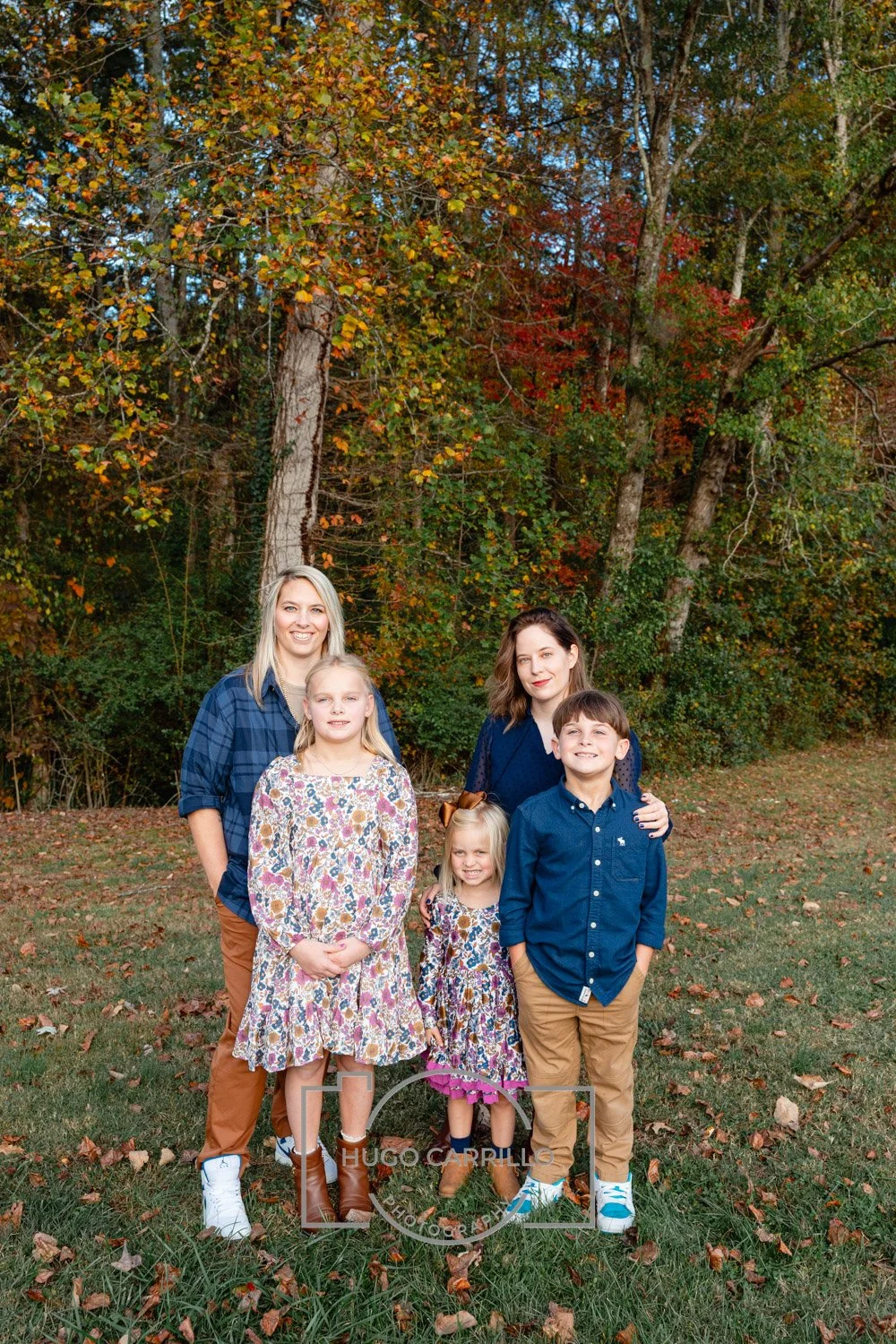 A family of five standing outdoors on a grassy area with autumn leaves, trees with fall foliage in the background.