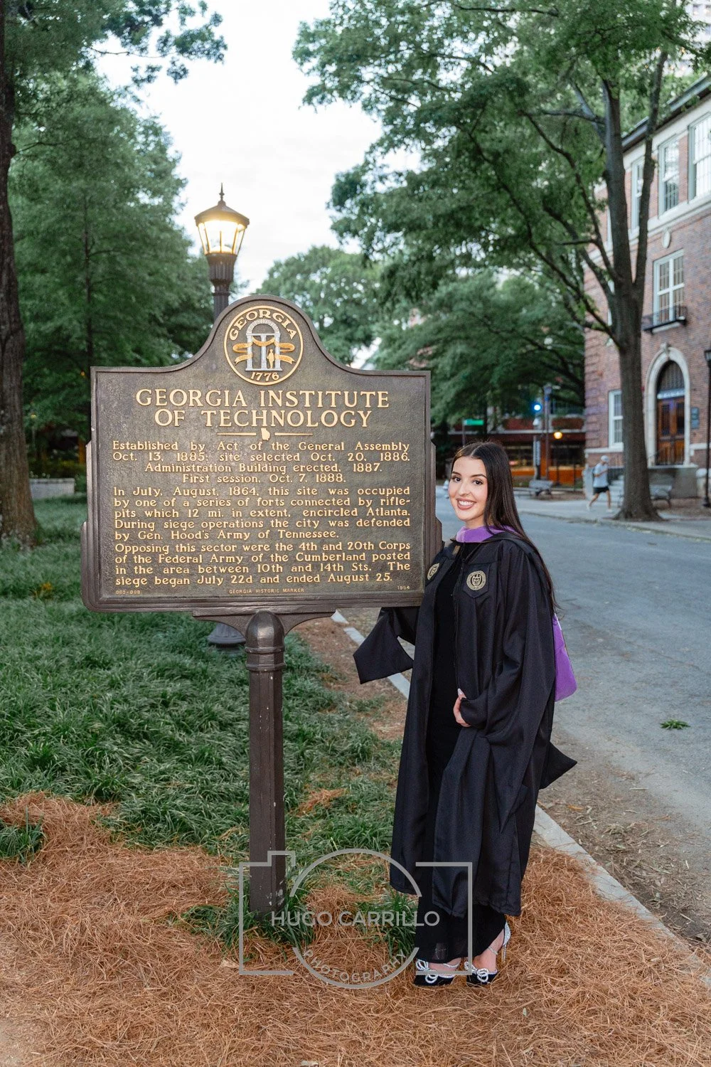 A woman in graduation gown and cap standing next to a historical marker for the Georgia Institute of Technology on a street with trees and buildings in the background.