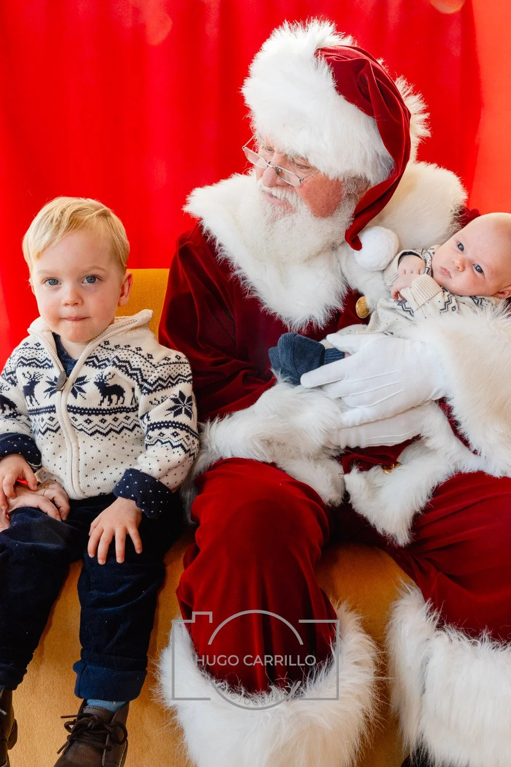A young blonde boy sitting next to Santa Claus dressed as Santa holding a baby, with a red backdrop.