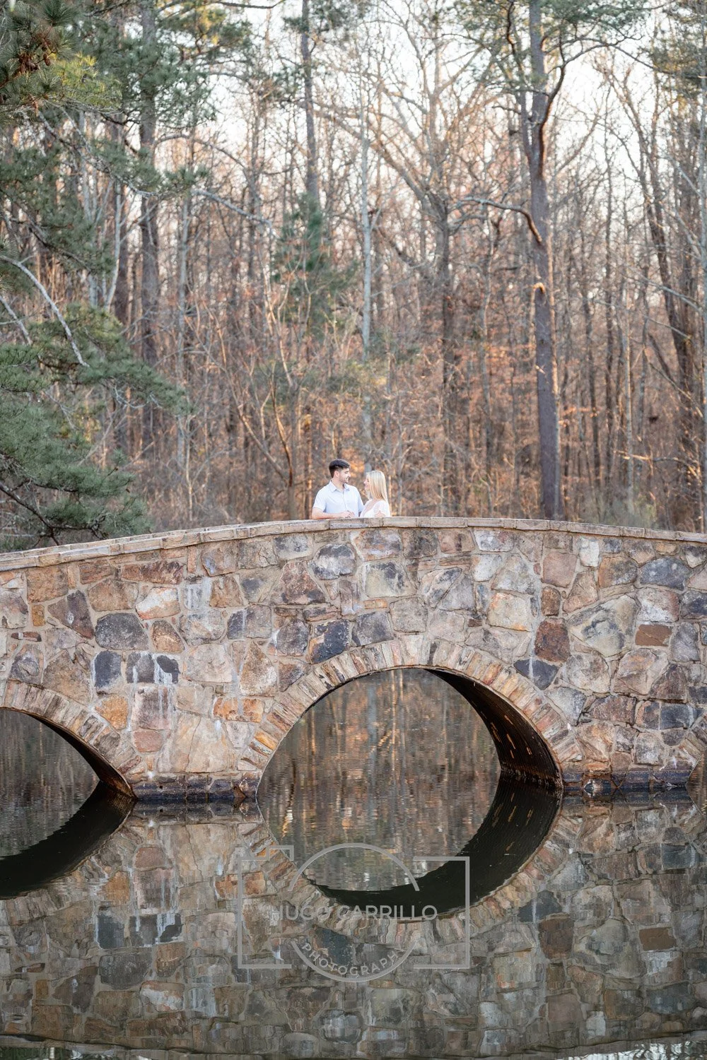 A couple standing on a stone bridge over a calm body of water, surrounded by tall trees with autumn foliage.