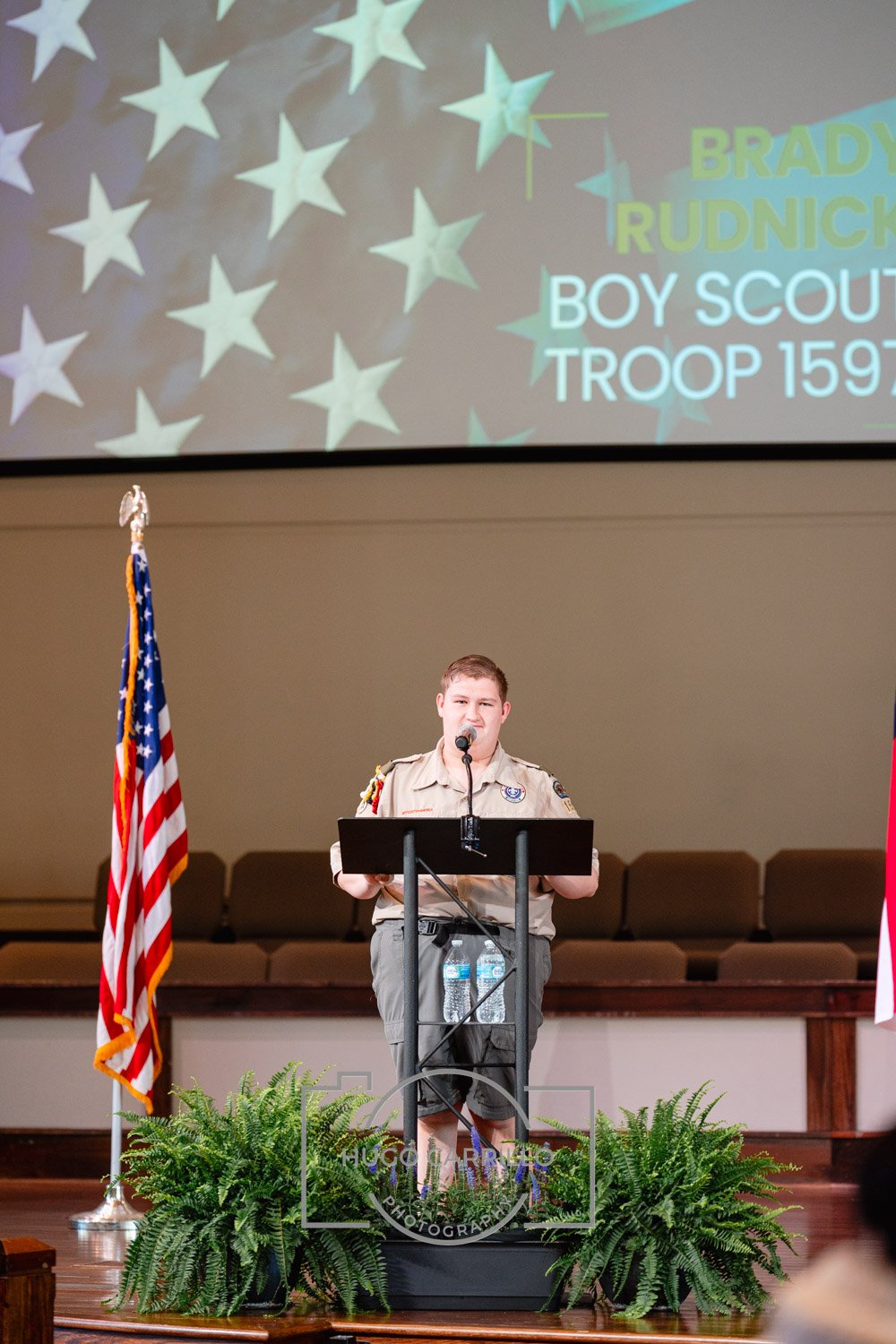 A young Boy Scout stands at a podium on a stage, speaking into a microphone. The stage is decorated with two American flags and green plants at the front. A large screen behind displays a blue star pattern and the text: 'BRADY RUDNICK BOY SCOUT TROOP