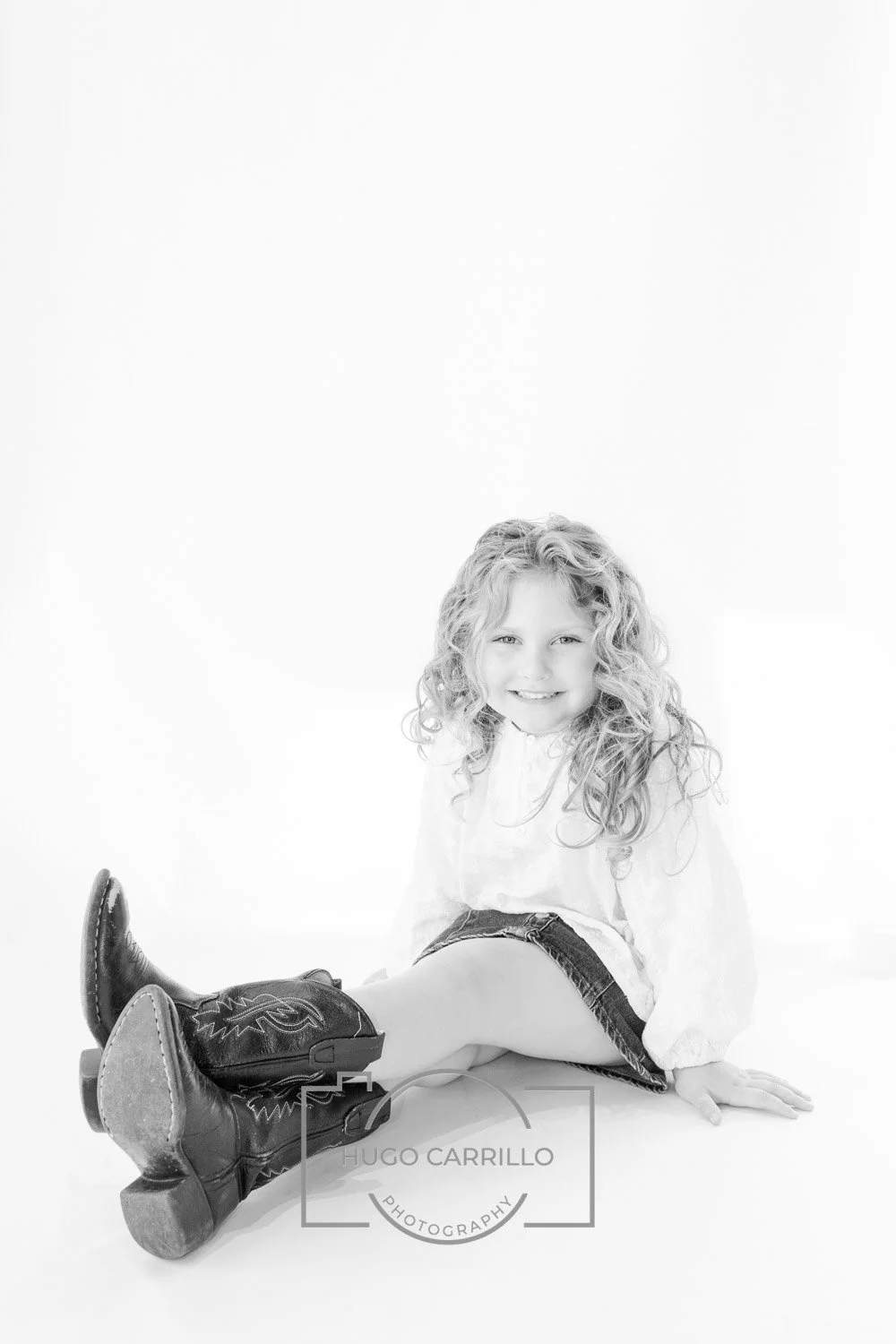 Smiling girl with curly hair sitting on the floor wearing cowboy boots, white blouse, and denim shorts, photography watermark at the bottom center.