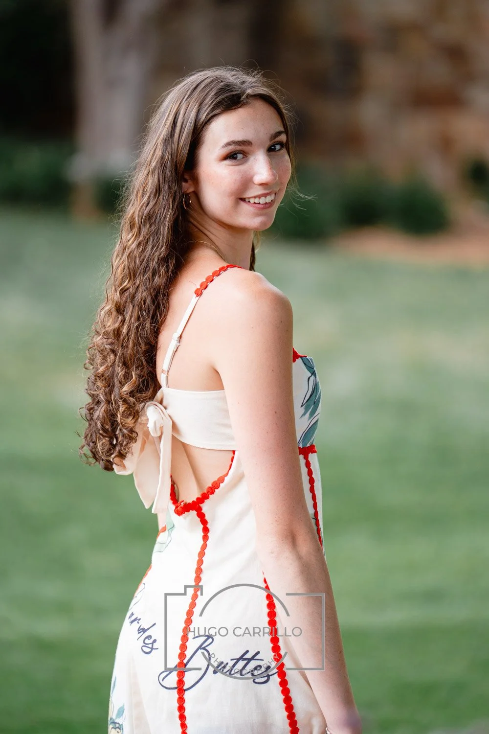 Young woman with long, curly brown hair, smiling, standing outdoors near a green field with trees in the background, wearing a cream-colored dress with orange accents and thin straps.