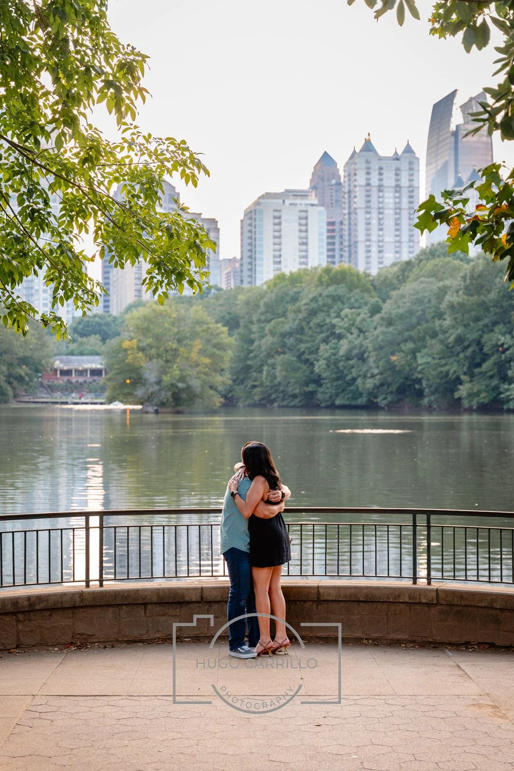 Two people hugging near a body of water with trees and city buildings in the background.