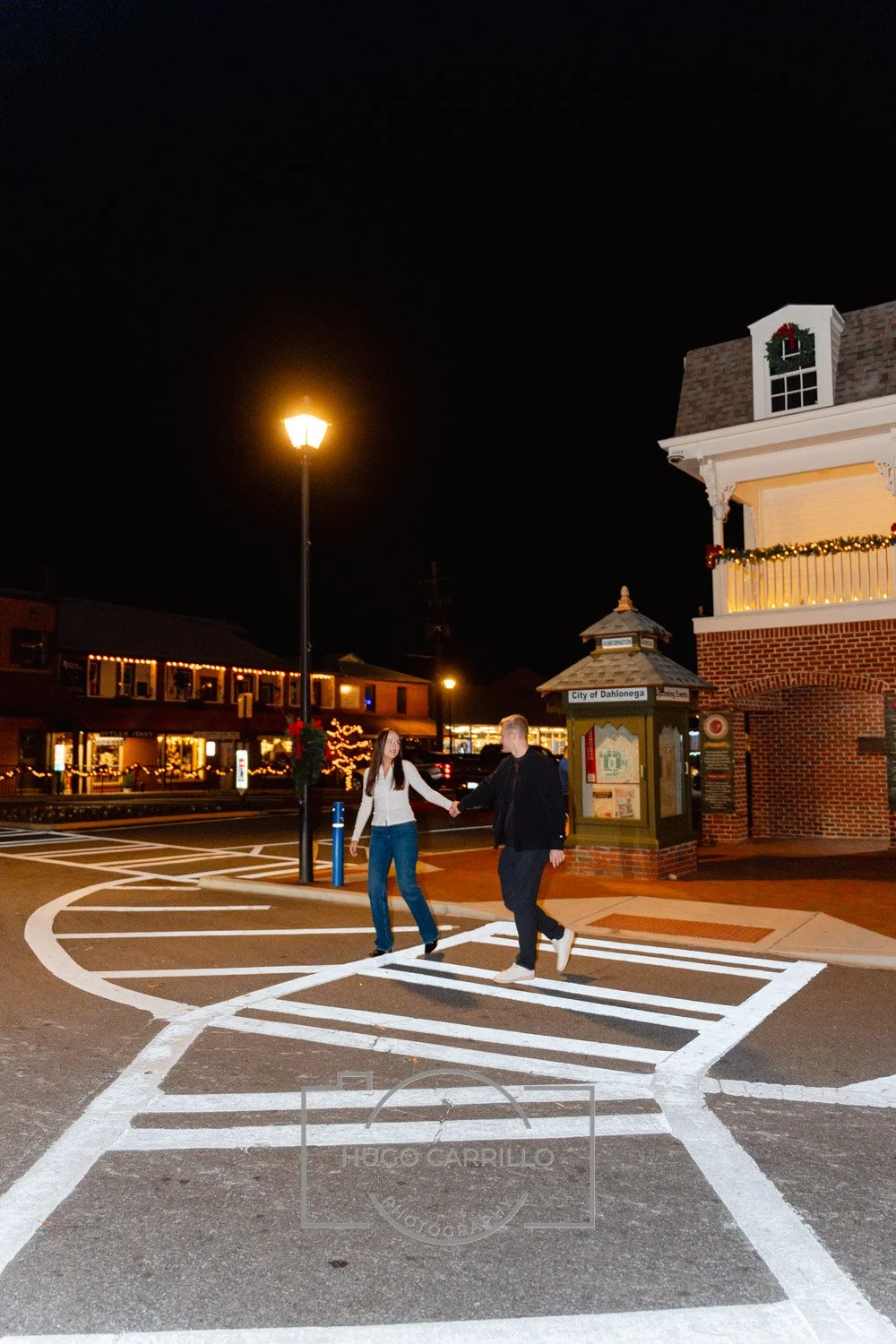 A young man and woman holding hands and crossing the street at night in a small town decorated for Christmas with Christmas lights, wreaths, and a Christmas tree in the background.