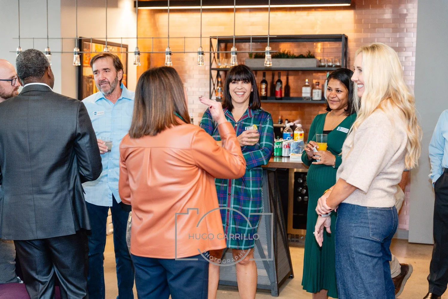 People socializing at a gathering in a modern kitchen with brick walls and bar shelves, holding drinks and engaging in conversation.