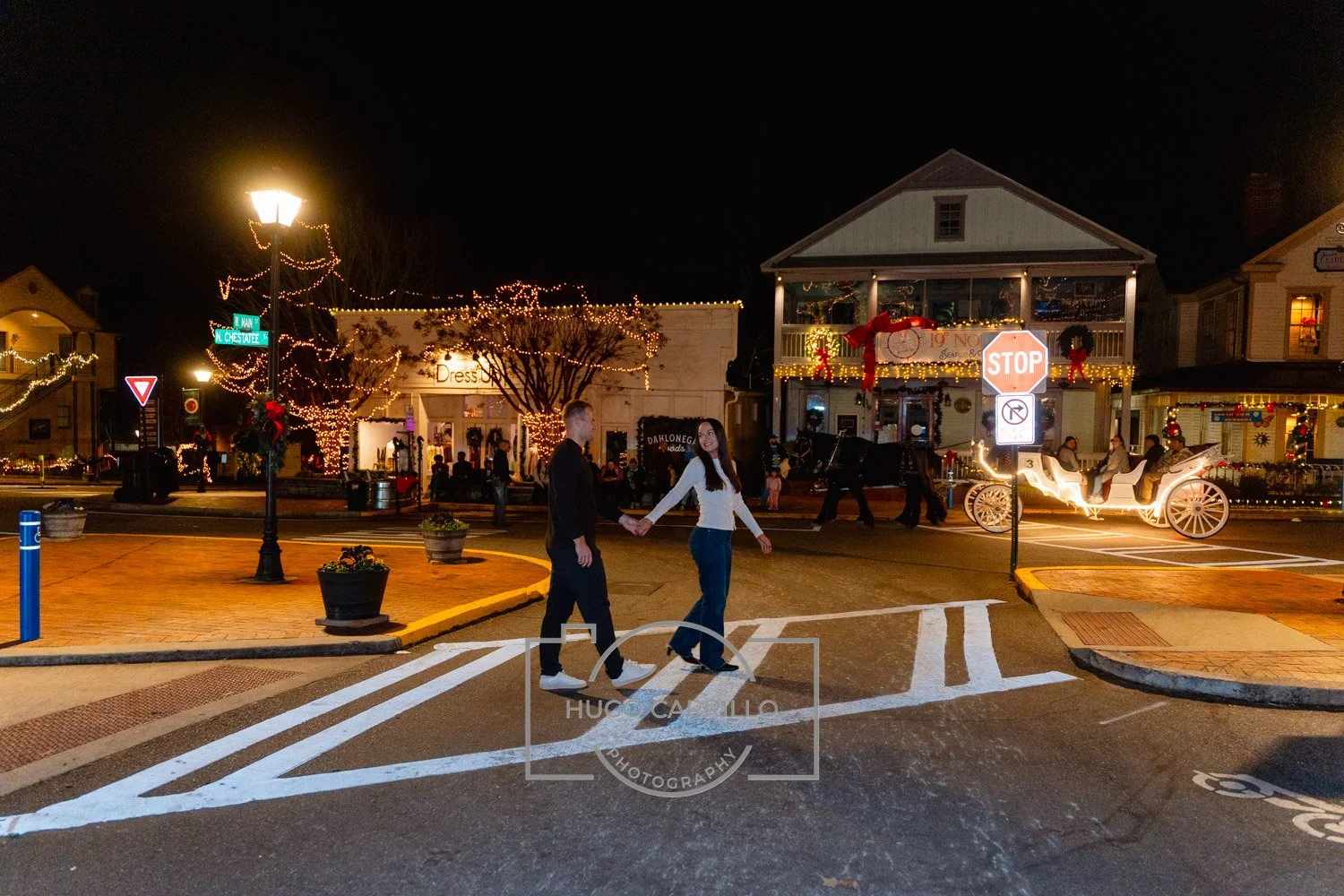 A couple holding hands and crossing the street at night with festive holiday decorations. There are string lights and wreaths on buildings, a horse-drawn carriage, and Christmas decorations on the storefronts.