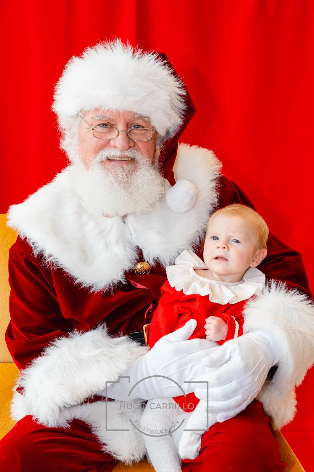 Santa Claus holding a small child in a red Christmas outfit with a white lace collar, sitting against a red background.