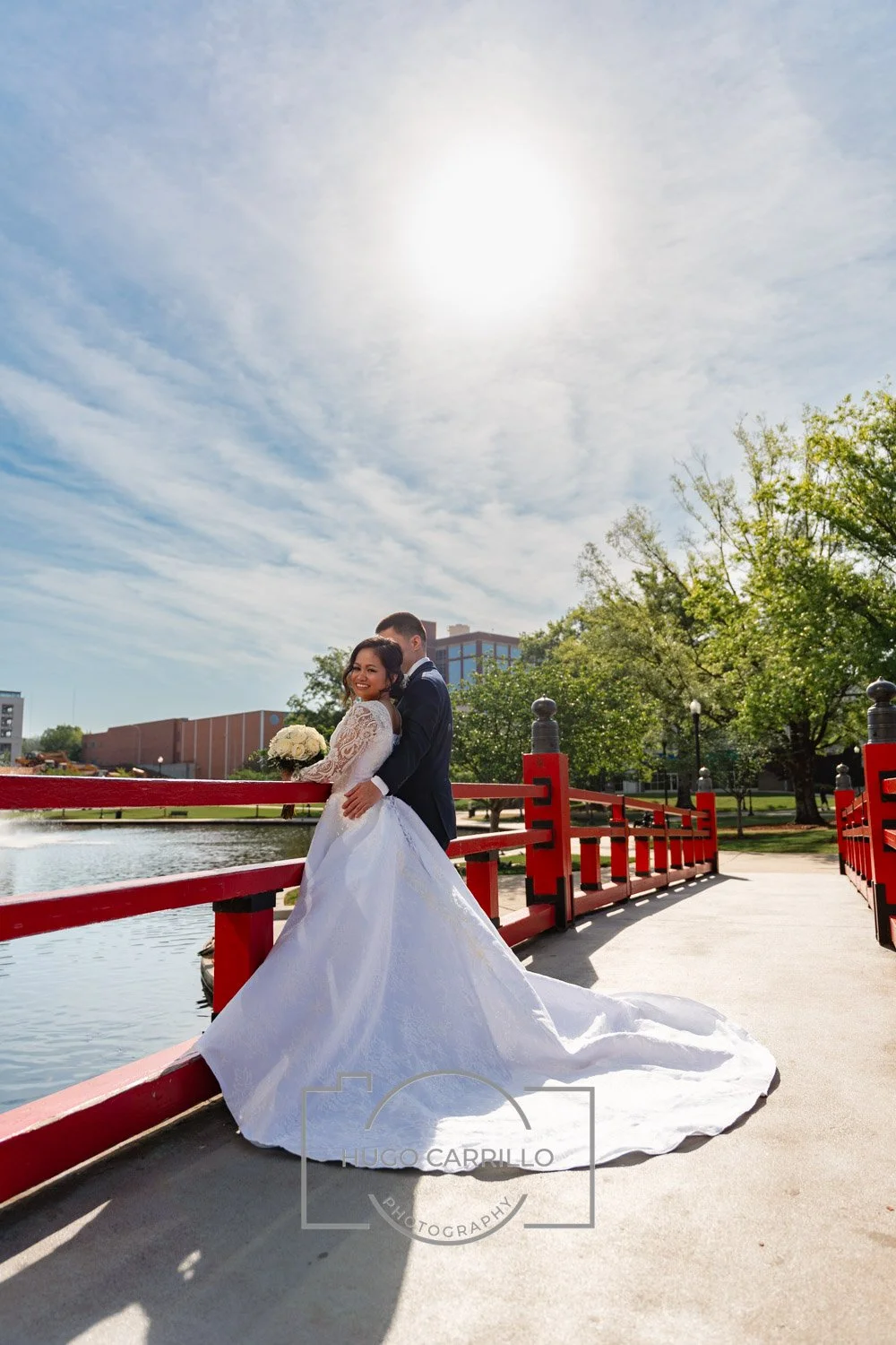 Bride in a white wedding gown holding a bouquet smiles as groom hugs her from behind on a red bridge by a pond in a park on a sunny day.