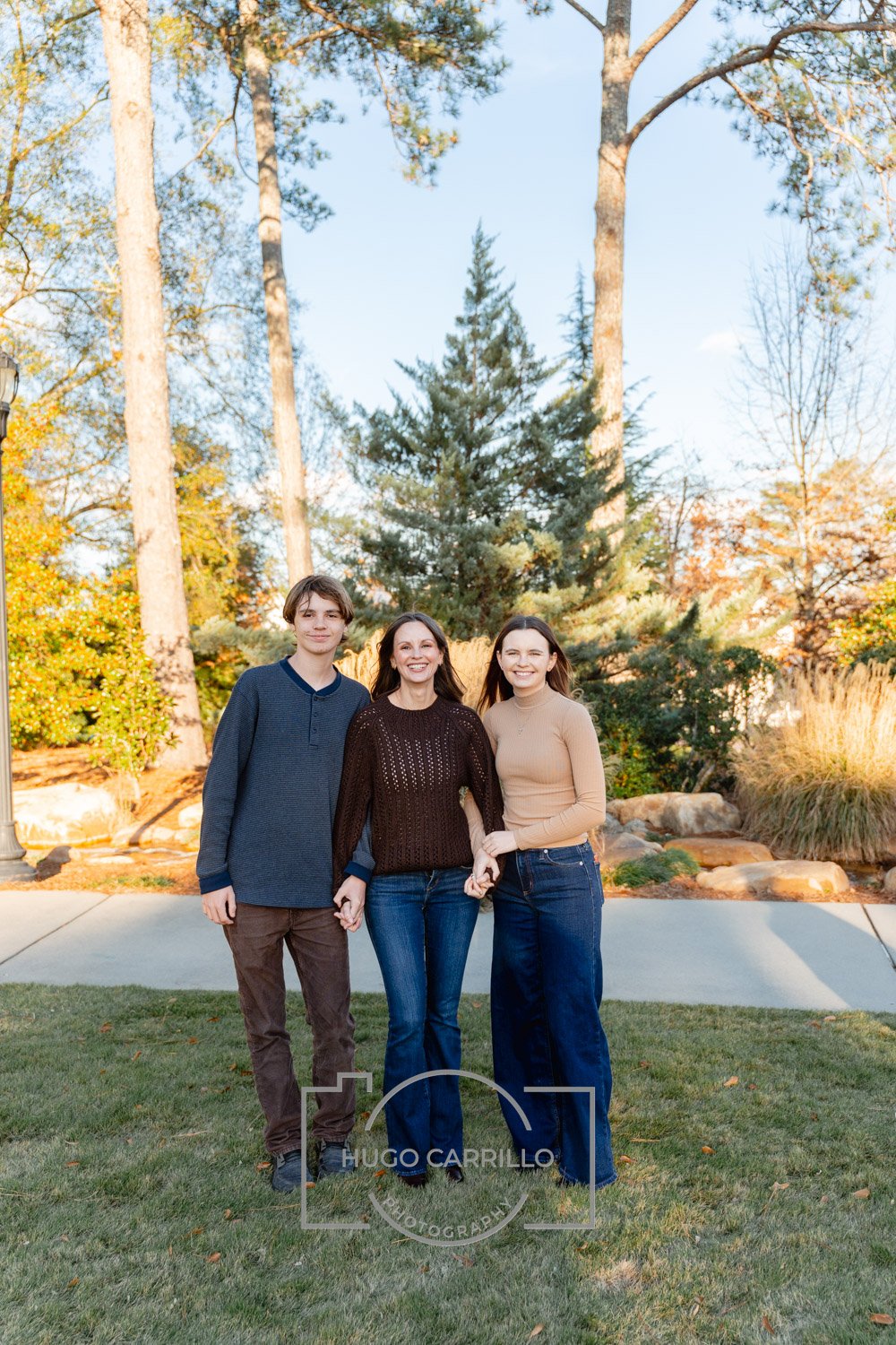 Three smiling people, two women and one young man, standing outdoors on a sidewalk with trees and autumn foliage in the background.